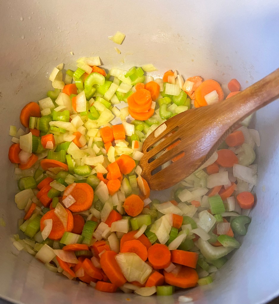 Chopped onions, carrots, and celery being sautéed in a pot with a wooden spoon, the base for a flavorful soup.