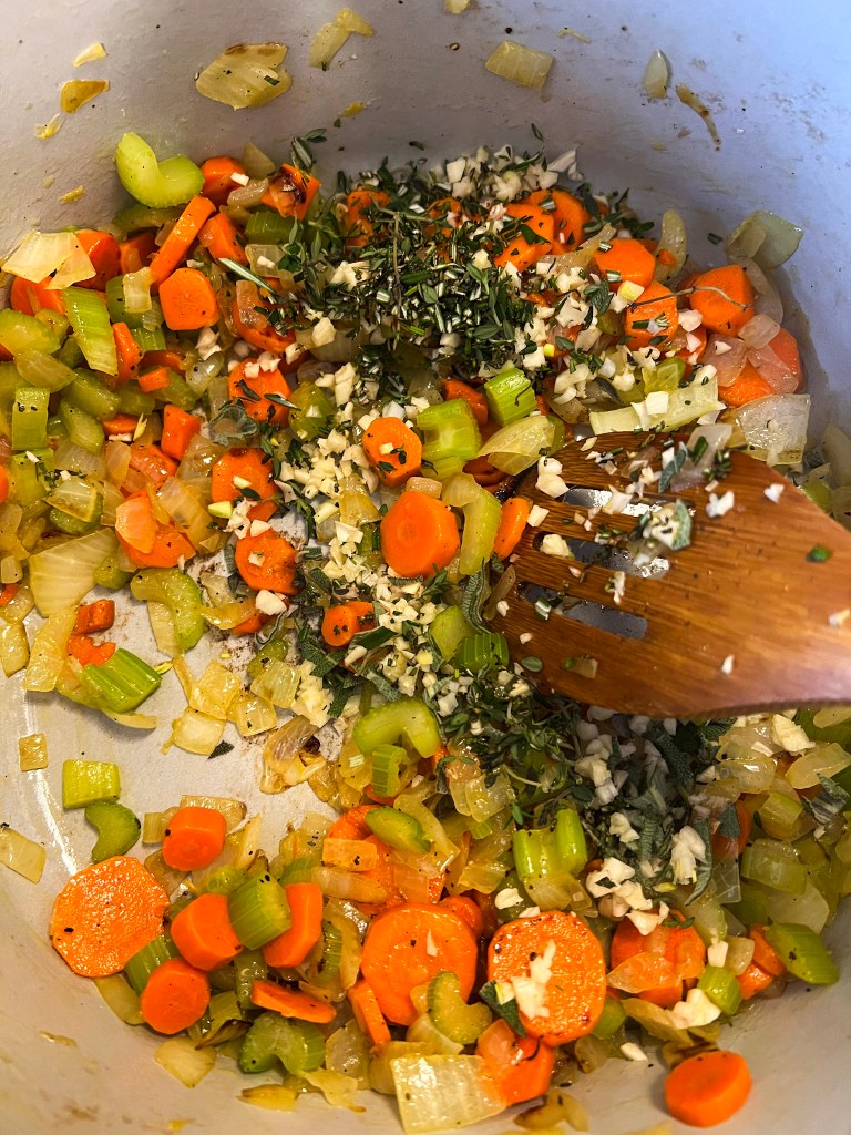 Sautéed carrots, celery, and onions in a pot, mixed with minced garlic and freshly chopped herbs, ready for soup preparation.