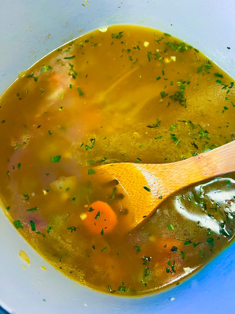 A wooden spoon stirs through a simmering pot of turkey soup with a clear broth, diced vegetables, and fresh herbs.