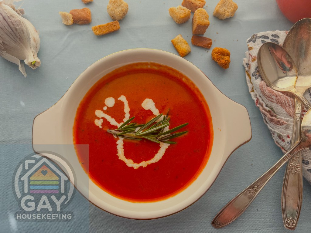 A bowl of vibrant tomato soup with a creamy swirl and rosemary garnish on a light blue tablecloth, next to a garlic bulb, croutons, a striped napkin, and silver spoons, with 'GAY Housekeeping' logo visible.